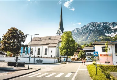 Eine malerische Kirche mit einem hohen Turm steht im Vordergrund. Im Hintergrund sind beeindruckende Berge und ein klarer Himmel zu sehen.