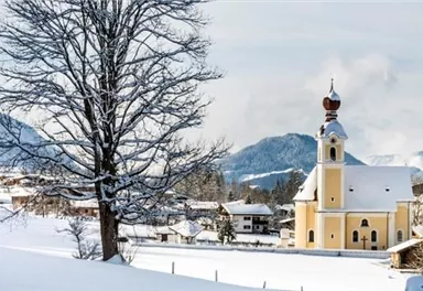 A picturesque snowy landscape with a yellow church and a large tree. The mountains in the background complete the wintry scene.