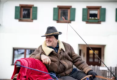 Ein lächelnder älterer Mann sitzt auf einer Kutsche. Im Hintergrund sieht man ein traditionelles Haus mit grünen Fensterläden.