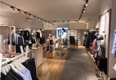 Ein moderner Bekleidungsladen mit gut sortierten Regalen und schlichten Möbeln. In der Mitte befindet sich ein Bildschirm, der Modepräsentationen zeigt.