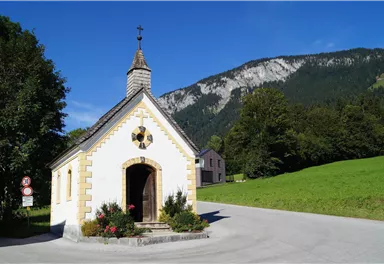 Eine kleine Kapelle steht an einer Straße in einer grünen Landschaft. Im Hintergrund sind Berge und ein blauer Himmel sichtbar.
