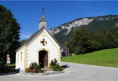 Eine kleine, charmante Kapelle steht an einer Kurve in einer malerischen Landschaft. Im Hintergrund sind grüne Wiesen und hohe Berge zu sehen.