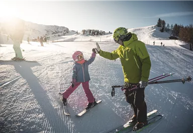 A father and his daughter are standing on skis in the snow. They are giving each other a high-five and enjoying a sunny day at the ski resort.