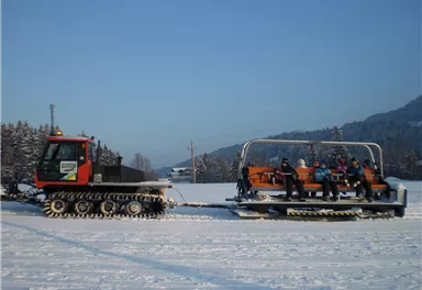Ein Schneemobil zieht einen Sitzanhänger mit mehreren Personen durch eine verschneite Landschaft. Die Umgebung ist winterlich und klar, mit Bergen im Hintergrund.