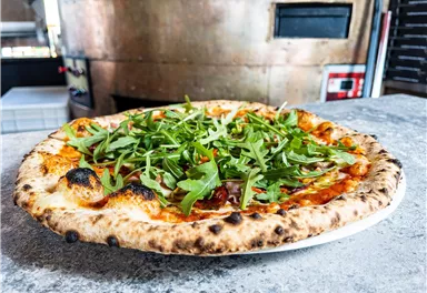 Eine frisch gebackene Pizza mit Rucola auf einem Teller. Im Hintergrund ist ein Pizzaofen sichtbar.