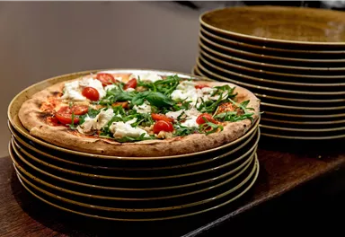 Eine frisch gebackene Pizza mit Tomaten und Rucola. Im Hintergrund sind mehrere Teller gestapelt.