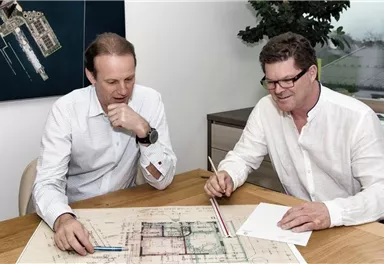 Zwei Männer sitzen an einem Tisch und betrachten einen Architektenplan. Sie diskutieren und machen Notizen auf Papier.