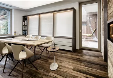 Ein modernes Esszimmer mit einem großen Tisch und weißen Stühlen. Durch die Glasfenster ist ein Blick nach draußen auf eine Terrasse sichtbar.