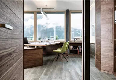 Ein modernes Büro mit großen Fenstern und Blick auf die Berge. Der Raum ist aus Holz gestaltet und enthält einen Schreibtisch sowie einen grünen Stuhl.