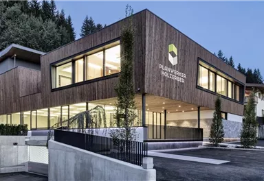 Ein modernes Gebäude mit Holzfassade und großen Fenstern. Es ist von Bäumen umgeben und wirkt einladend.