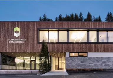 Ein modernes Gebäude mit Holzverkleidung und großen Fenstern. Im Vordergrund befindet sich eine grüne Pflanze und im Hintergrund sind Bäume zu sehen.