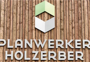 Ein Holzschild mit dem Schriftzug "PLANWERKER HOLZERBERG". Darüber befindet sich ein geometrisches Logo in Grün und Weiß.