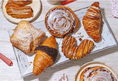 Eine Vielzahl von köstlichen Gebäckstücken auf einem Holzbrett. Darunter befinden sich Croissants, Zimtschnecken und gefüllte Teilchen.