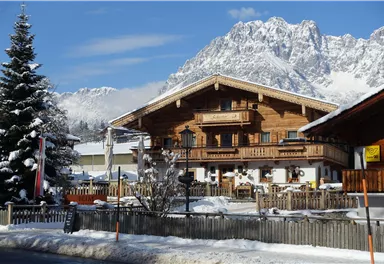 A beautiful wooden house in winter with a snow-covered roof and a picturesque mountain landscape in the background. The sky is clear and blue.