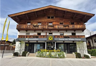 Eine moderne Bankfiliale im alpinen Stil mit Holzverkleidung. Vor der Bank stehen gelbe Fahnen und es gibt eine grüne Bepflanzung.