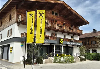 Ein modernes Gebäude im alpinen Stil mit der Aufschrift „Raiffeisen Immobilien“. Vor dem Gebäude stehen gelbe Fahnen und es gibt einen klaren Himmel im Hintergrund.