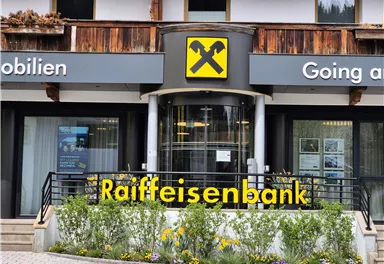 Die Fassade der Raiffeisenbank mit einem modernen Eingang und grünen Pflanzen vor dem Gebäude. Das Schild zeigt deutlich den Namen der Bank.