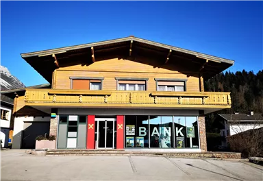 A modern bank building in alpine style with wooden cladding. In the foreground, large window panes with the inscription "BANK" are visible.