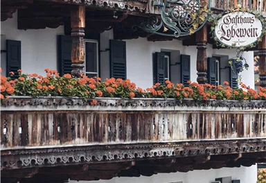 Ein traditionelles Gasthaus mit einem hölzernen Balkon, der mit bunten Blumen geschmückt ist. Die Fassade ist im Alpenstil gestaltet, mit Fenstern und einer einladenden Eingangstür.