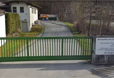 Ein grünes Tor an einem schmalen Weg, umgeben von Häusern und Bäumen. Im Hintergrund ist eine Straße sichtbar.