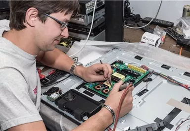 A man is working on an electronic device and inspecting the circuit board. Tools and parts are around him on a table.