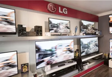 A modern electronics store with several LG televisions displayed in a row on shelves. Various devices like speakers and Blu-ray players are also visible.