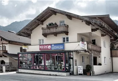 A traditional building with a shop on the ground floor and balconies on the upper floor. The surroundings showcase rural architecture and a mountainous backdrop.