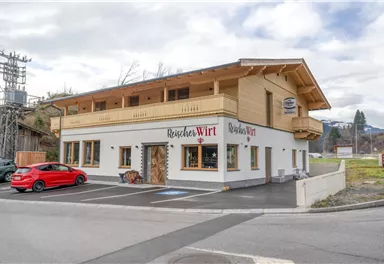 Ein modernes Gebäude im alpinen Stil steht an einer Straßenecke. Vor dem Haus parkt ein rotes Auto.