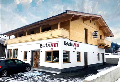 A modern building with wooden cladding and a bright outdoor area. The inscription "Reischer Wirt" is clearly visible on the facade.