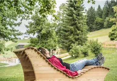 A person is lying relaxed on a wooden lounger in the midst of a green landscape. In the background, trees and a gentle hilly landscape can be seen.