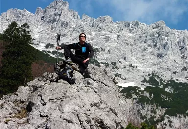 A hiker is sitting on a rock and enjoying the view of snow-capped mountains. The sky is blue with a few clouds.
