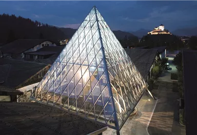 Eine moderne Glaspyramide steht im Vordergrund, umgeben von Gebäuden. Im Hintergrund ist eine Burg auf einem Hügel zu sehen.