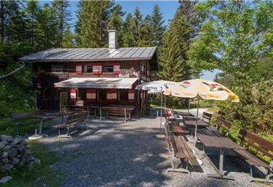 Eine gemütliche Berghütte umgeben von Bäumen. Vor der Hütte stehen Bänke und Tische, ideal für eine Pause in der Natur.