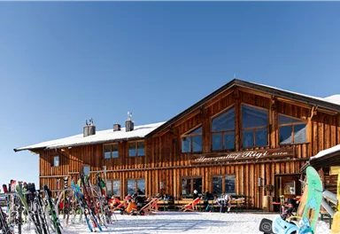 Eine gemütliche Berghütte aus Holz in schneebedeckter Landschaft. Vor der Hütte stehen Skier und Snowboards.