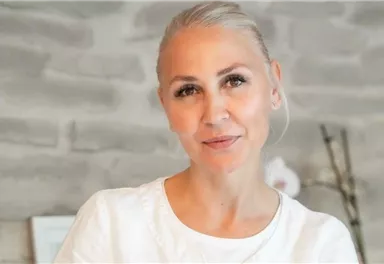 A woman with blonde hair and a friendly smile is standing in front of a gray wall. She is wearing a simple white top and looks relaxed.
