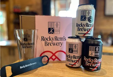 Eine Sammlung von Bierdosen und einem Glas der Brauerei Rocky Rem's. Das Ensemble befindet sich auf einem Holztisch und zeigt die typischen Markenfarben und das Logo.