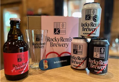 Eine Arrangement von Getränken und Zubehör von Rocky Rem's Brewery. Dazu gehören eine Flasche Bier, Dosen, ein Glas und ein Flaschenöffner.