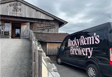 Ein Holzgebäude mit dem Schriftzug "Mahlberg Rem" und ein schwarzes Lieferfahrzeug mit der Aufschrift "Rocky Rem's Brewery". Die Szene zeigt eine ländliche Umgebung mit grauem Himmel.