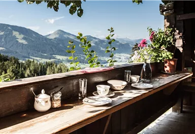 A beautiful table with dishes and glasses on a wooden terrace. In the background, mountains and a clear, sunny view can be seen.