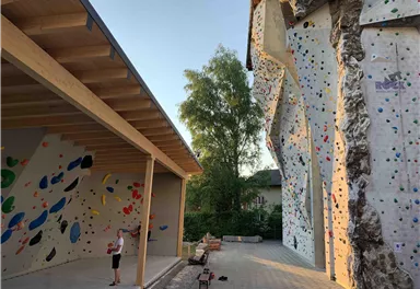An outdoor climbing wall with colorful grips and a climber who is training. In the background, trees and a second climbing wall are visible.