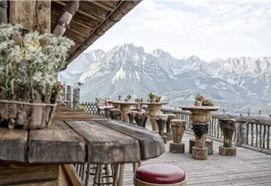 Eine Holzterrasse mit rustikalen Tischen und Blumen. Im Hintergrund erheben sich beeindruckende Berge unter einem klaren Himmel.