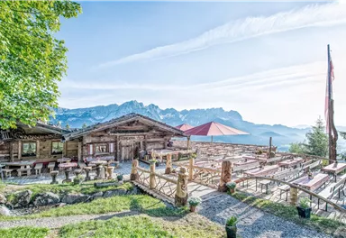 Eine gemütliche Berghütte mit traditionellem Holzdesign und einer schönen Terrasse. Im Hintergrund sind majestätische Berge und ein blauer Himmel zu sehen.