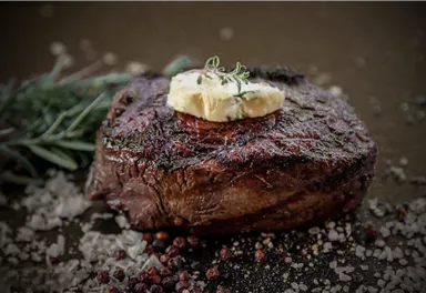 A juicy steak, perfectly grilled and garnished with herb butter. Surrounding it are some spices and fresh herbs.