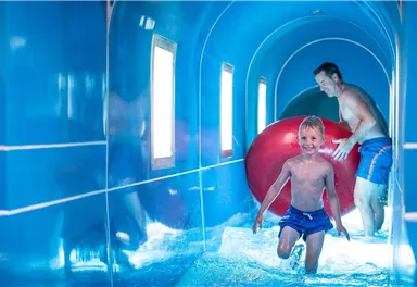 Ein Junge läuft fröhlich in einem blauen Wassertunnel, während ein Erwachsener ihm mit einem großen roten Ball folgt. Die Atmosphäre ist lebhaft und einladend.