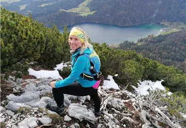 An active person is hiking up a steep mountain. In the background, there is a beautiful lake and forested mountains.