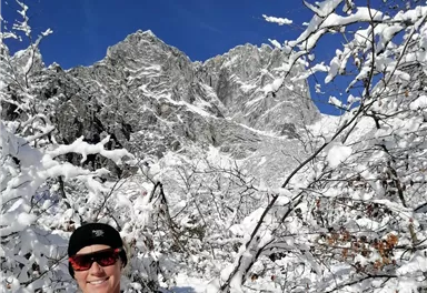 A person is standing in a snow-covered forest with mountains in the background. The sky is clear and blue.