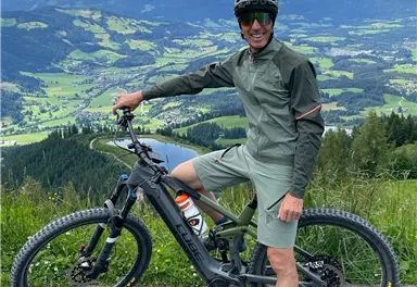A mountain biker proudly stands next to his e-bike in the mountains. In the background, green valleys and majestic mountains are visible.