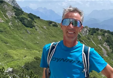 A man is standing smiling in nature on a mountain. In the background, green meadows and mountainous landscapes can be seen.