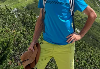 A man stands on a mountain path wearing sporty clothing. In the background, green hills and a clear blue sky can be seen.