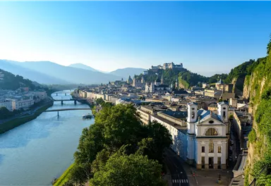A picturesque view of the city of Salzburg with the Salzach River in the foreground. In the background, mountains and historical buildings stretch out.